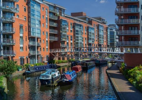 The canal district in Birmingham, with high rise buildings either side of the canal and colourful boats passing beneath the bridge.
